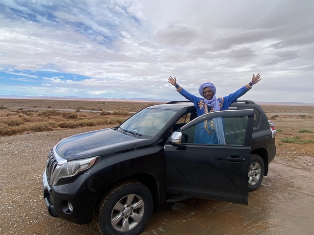 Person in traditional clothing standing by a car in the desert.