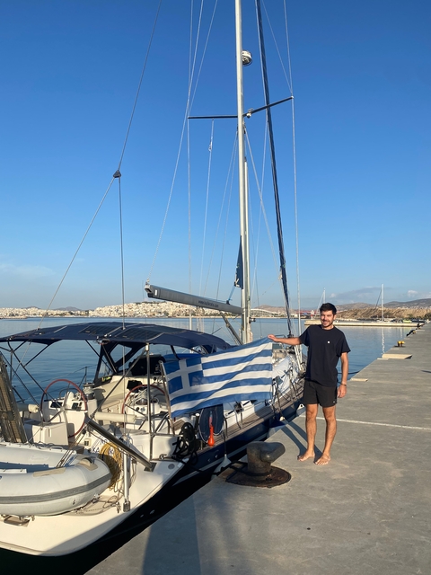 Person holding Greek flag next to a sailboat at a marina.