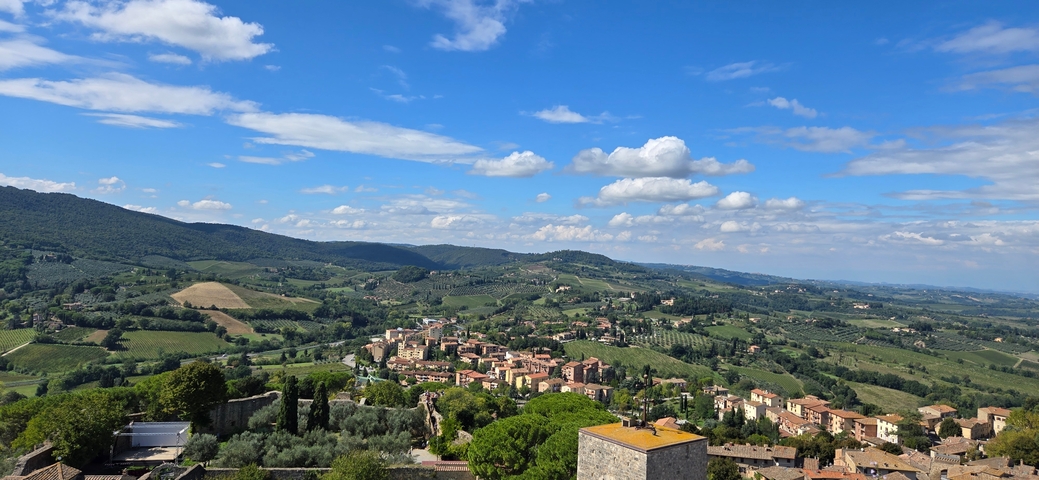       A scenic landscape view of a countryside with a village surrounded by lush greenery and rolling hills under a blue sky with clouds.
  