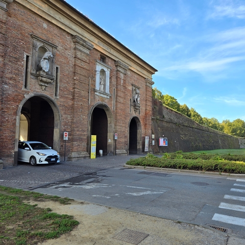       A medieval gateway with arches and a modern car passing through, set against a backdrop of greenery.
  