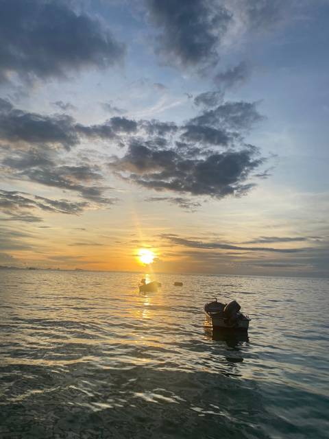       A lone boat drifts on calm tropical seas beneath a vibrant orange sunset and dramatic clouds.
  
