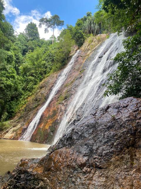       A tall jungle waterfall flows over streaked rock into dense green vegetation.
  