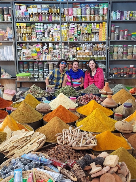       Three people sitting in front of a variety of spices in a market.
  