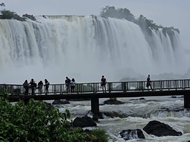 People walking on a viewing platform in front of a large waterfall.