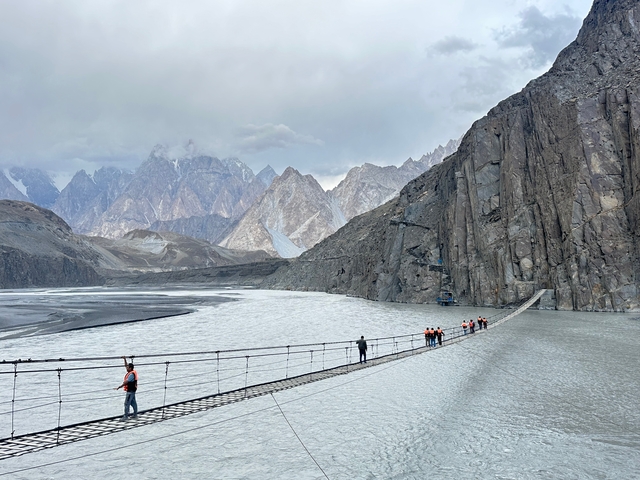 People crossing a suspension bridge over a river with mountains