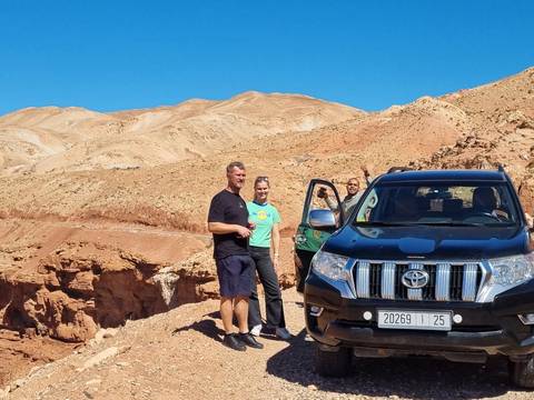       Three people standing beside a vehicle with desert hills in the background.
  