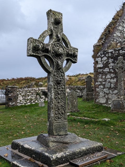 A historic stone Celtic cross in an ancient graveyard with a cloudy sky.