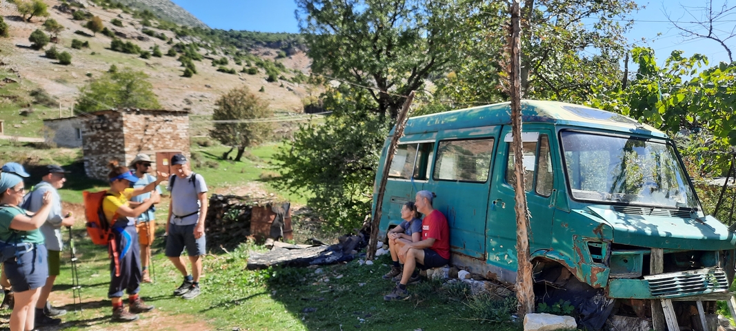       Hikers resting beside an old, worn-out vehicle in a rural setting.
  
