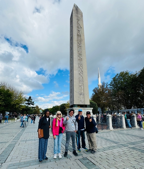 Group of people posing in front of a historical obelisk with a mosque visible.