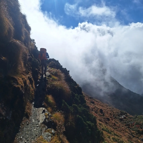       Hiker walking along a narrow mountain path under cloudy skies.
  