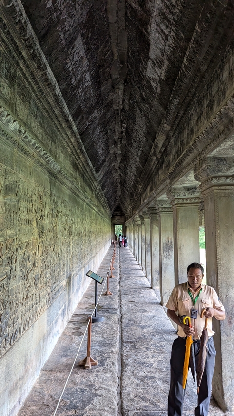       People walking along an ancient temple corridor with intricate carvings.
  