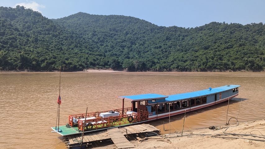 Long boat moored by the riverside with wooded hills in the background.