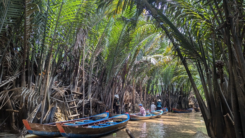       People on traditional boats navigating a narrow river through dense greenery.
  