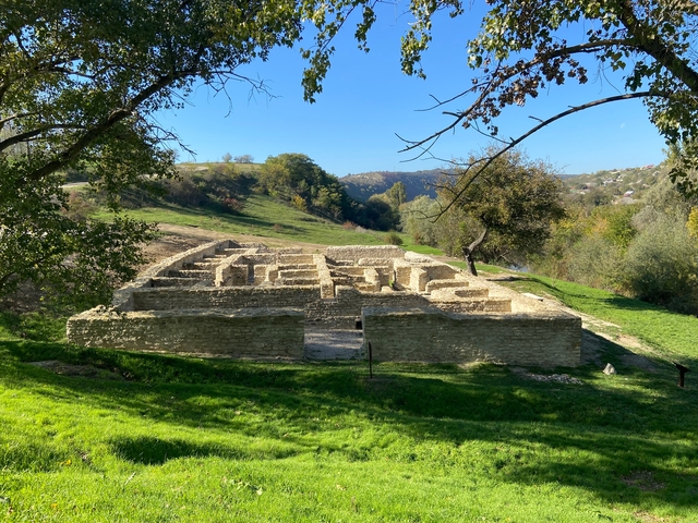       Archaeological ruins in a green hilly landscape under clear blue skies.
  