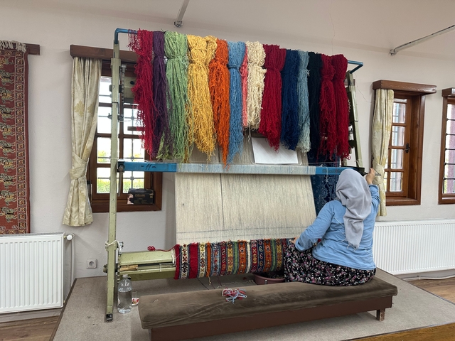       Woman working on a traditional carpet loom with colorful threads.
  
