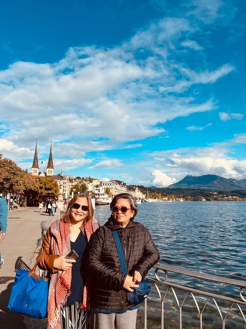       Two people posing by a lake with mountains and a cityscape.
  
