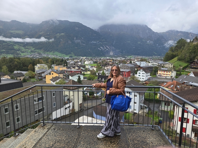       Person posing on a balcony with a view of a town set in a mountainous area.
  