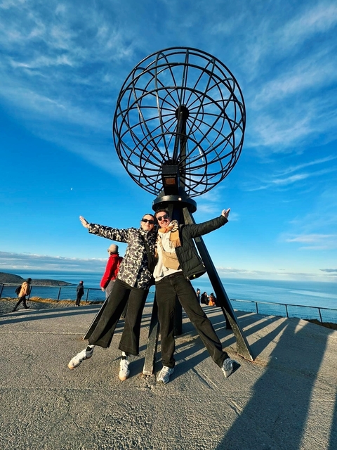       Two people posing under a globe sculpture with coastline in the background.
  