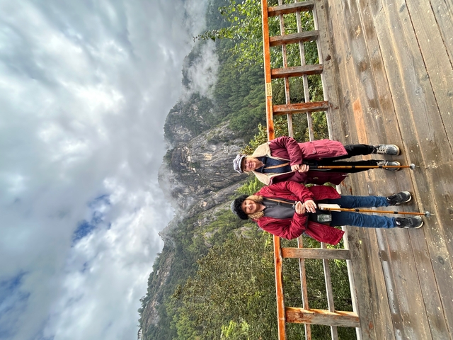      Two people with hiking sticks on a wooden path overlooking mountains
  
