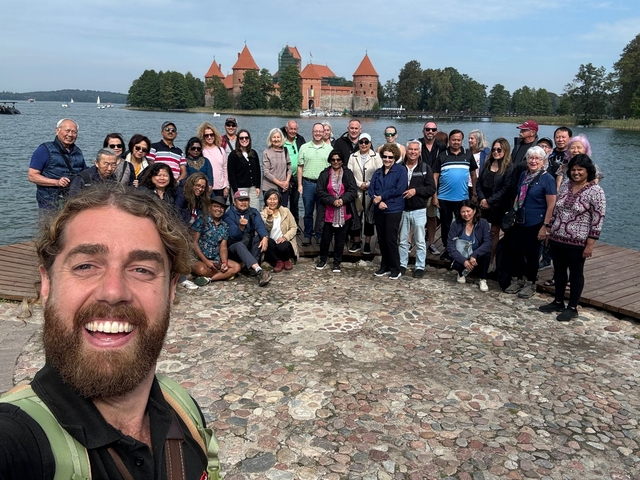 A large group of people posing for a photo by a lake with a castle in the background.