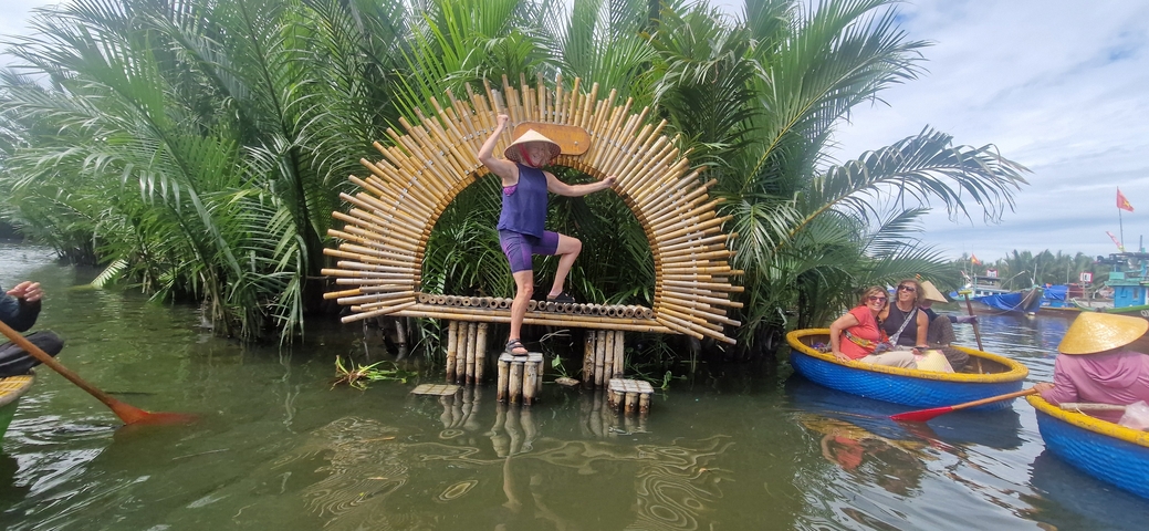       Person posing on a bamboo structure in a river, with a traditional boat nearby.
  