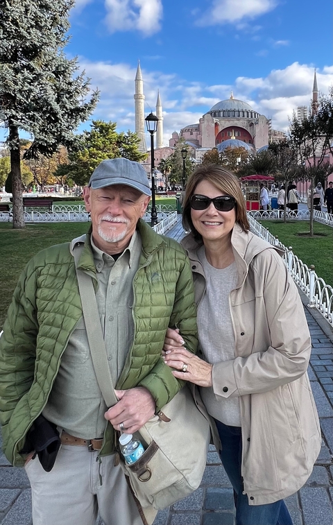 A couple posing in front of a park with people in the background.