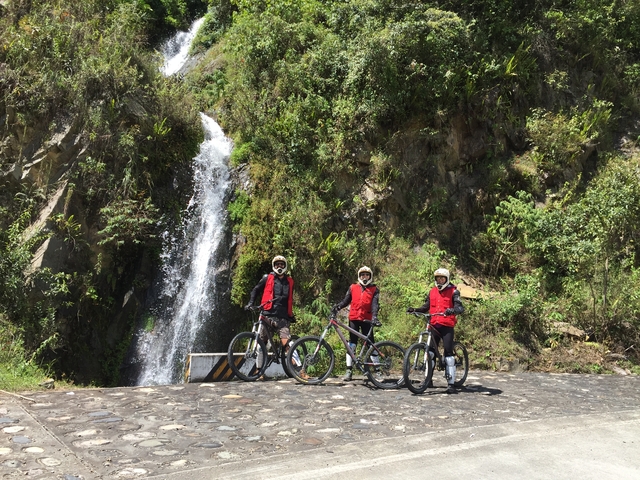       Cyclists wearing helmets posing in front of a waterfall.
  