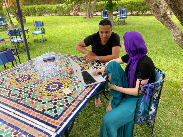       Two people working on a laptop at a mosaic table outdoors.
  