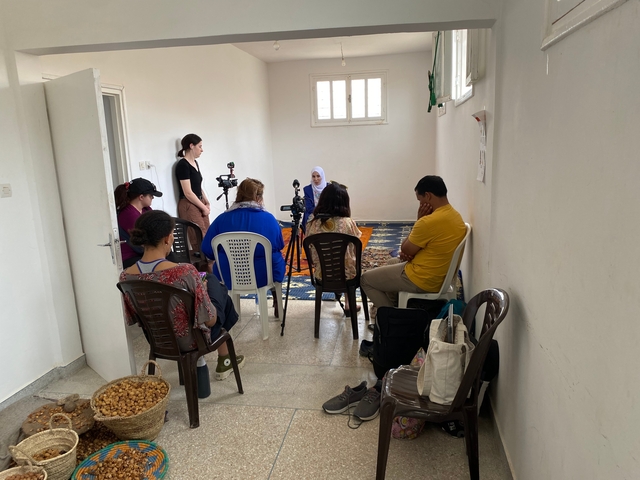       People seated in chairs, facing a person speaking to a camera in a bright room.
  