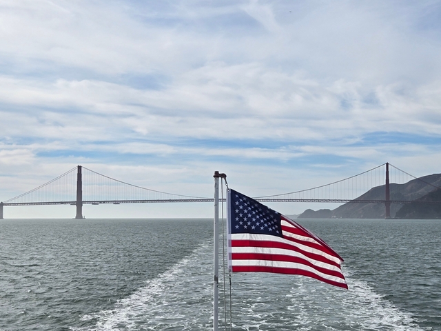 Golden Gate Bridge under a bright sky with American flag in foreground.