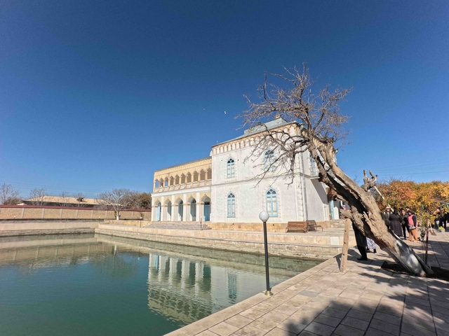       Building by a reflective pool with a tree and visitors around.
  