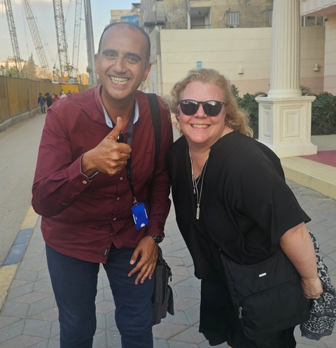       Two people posing next to a street with one giving a thumbs up.
  