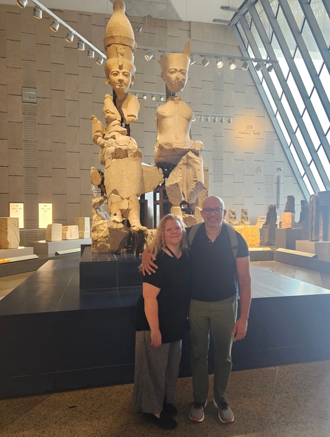       Two people smiling and posing inside a museum with large stone figures.
  