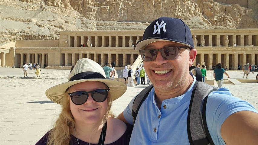       A smiling couple taking a selfie in front of a large ancient structure.
  