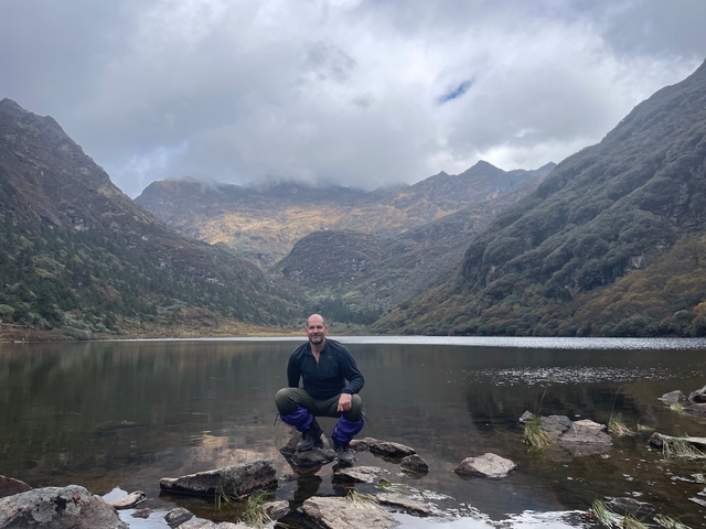 Person posing in front of a lake surrounded by mountains.