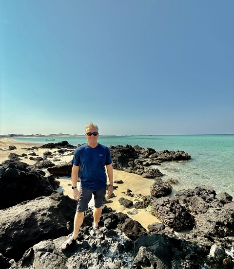 Man posing on a rocky beach with clear blue water.