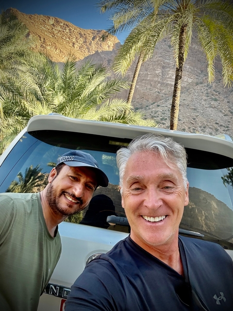 Two men posing with a car window backdrop of trees and mountains.