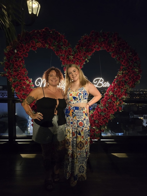 Two women posing in front of a heart-shaped floral display at a bar