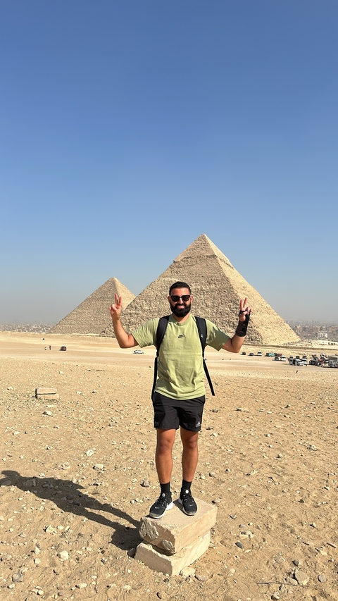 Person posing with pyramids in the background on a clear day.