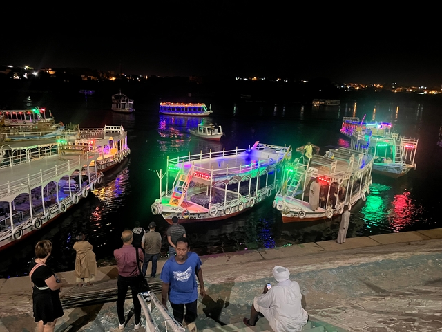       Boats on a river with colorful lights at night, people on the bank.
  