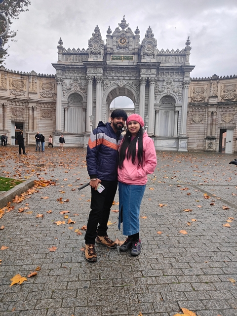 Couple posing in front of an ornate gate with stone carvings and autumn leaves on the ground.