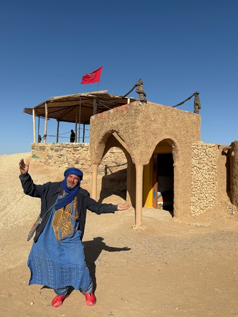       Man posing joyfully in front of a mud-brick structure.
  