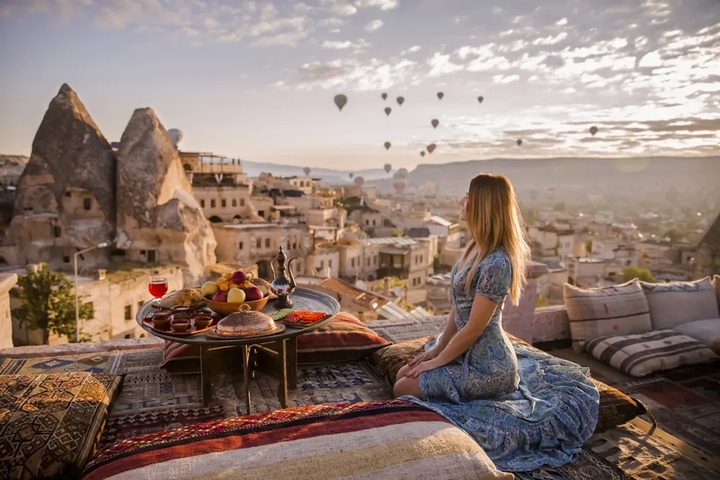 A woman enjoying breakfast on a rooftop terrace with a view of Cappadocia and hot air balloons.