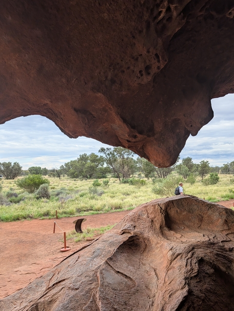 Person walking under a large rock overhang, surrounded by grass.
