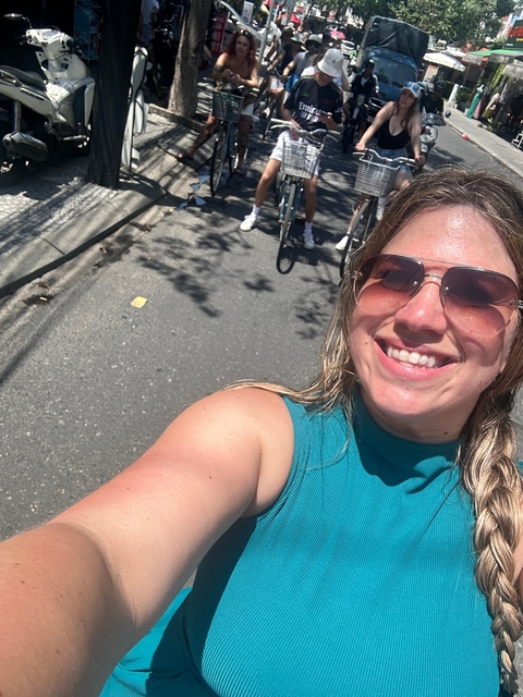 A close-up of a person smiling on a street with cyclists in the background.