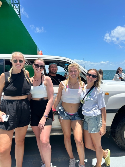       A group of people standing by a white vehicle, smiling on a sunny day.
  