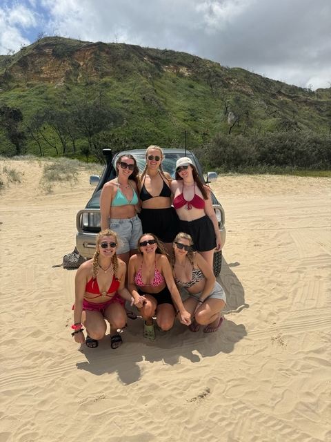 Six women posing on a sandy beach in front of a vehicle.