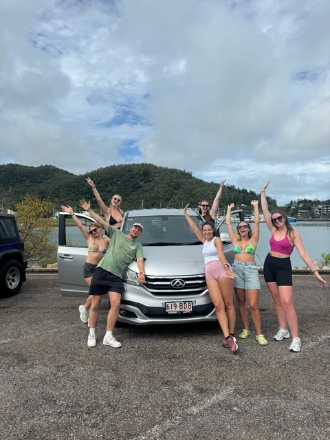 A group of people posing in front of a silver van with raised arms, with hills in the background.