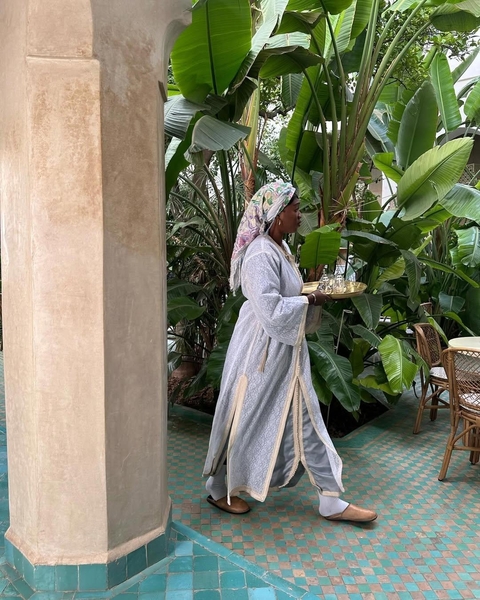 Woman in traditional clothing serving tea in a lush courtyard.