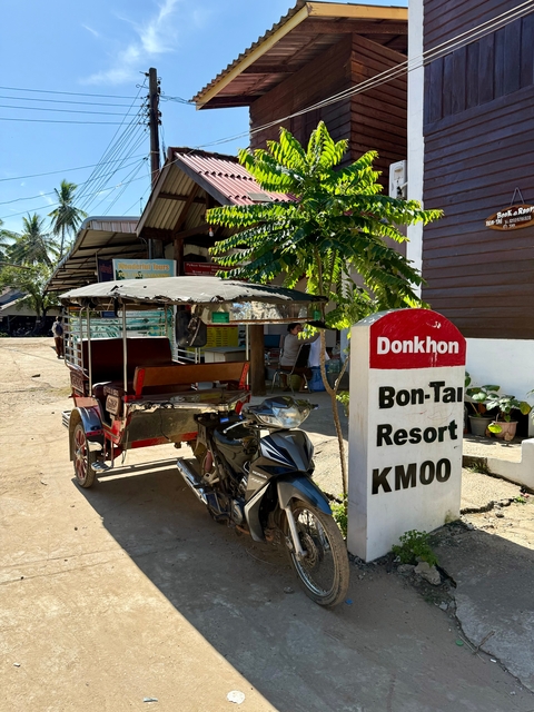 Colorful tuk-tuk near a resort sign next to leafy trees.